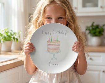 May include: A round white ceramic plate featuring a watercolour illustration of a layered cake on a light blue pedestal stand. The text Happy Birthday is printed above the cake, and the name Ciera is printed below in a simple black font.