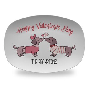 May include: An oval white serving platter featuring a graphic of two brown dachshunds wearing sweaters and kissing. Above the dogs is the red text Happy Valentine's Day, and below them is the black text THE FRAMPTONS with two small hearts.