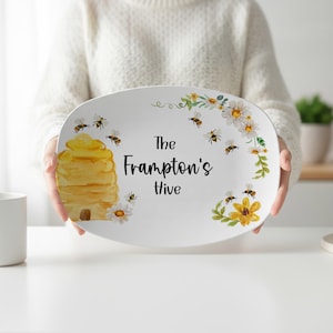 May include: An oval white serving platter featuring a watercolor illustration of a yellow beehive, several small bees, and white and yellow daisies. The text "The Frampton's Hive" is printed in black script and sans-serif lettering in the center.
