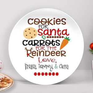 May include: A round white ceramic plate featuring the text Cookies for Santa and Carrots for the Reindeer in brown, red, and black lettering. The design includes a tan cookie, an orange carrot, and the names Briana, Tommy & Ciepa in black.