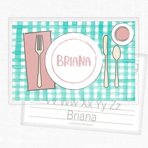 May include: A rectangular placemat featuring a teal and white watercolor gingham pattern. It displays a pink napkin, a fork, a knife, a spoon, and a small bowl. A central white plate features the name BRIANA in pink text. Below, a practice sheet shows the alphabet and the name Briana.