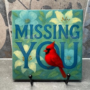 May include: A blue and green ceramic tile with a white flower and a red cardinal bird. The tile has the words "MISSING YOU" in blue letters.