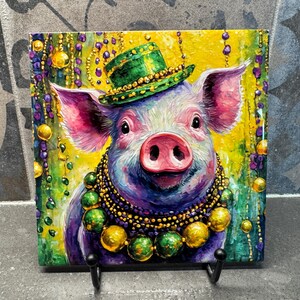 May include: A square ceramic tile featuring a whimsical illustration of a pig wearing a green top hat and a beaded necklace. The pig is painted in vibrant colors, with a yellow background and Mardi Gras-themed beads.
