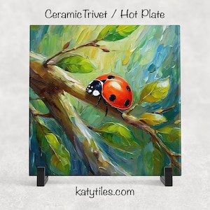 May include: Square ceramic trivet with a painting of a ladybug on a branch with green leaves. The ladybug is red with black spots. The trivet has the text "Ceramic Trivet / Hot Plate" and "katytiles.com".