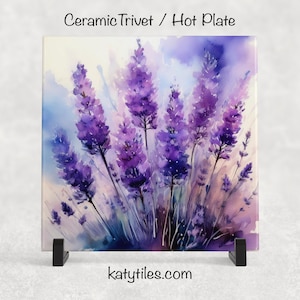 May include: Square ceramic trivet with a watercolor painting of lavender flowers in shades of purple and white. The trivet is displayed on a black stand. Text at the top reads "Ceramic Trivet / Hot Plate" and the website "katytiles.com" is at the bottom.