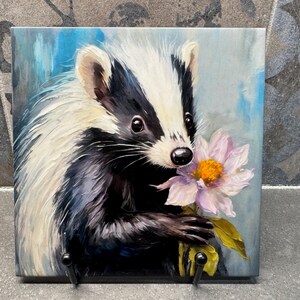 May include: Square ceramic tile featuring a badger holding a pink flower. The badger has black and white fur with a detailed painting style. The background is a blend of blue and white brushstrokes. Two black hooks are attached to the bottom.