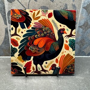 Puede incluir: Arte mural cuadrado con una colorida ilustración de un pájaro con vibrantes plumas naranjas, rojas y azules. La obra de arte incluye un fondo crema con detalles de hojas y flores, y dos ganchos negros en la parte inferior.