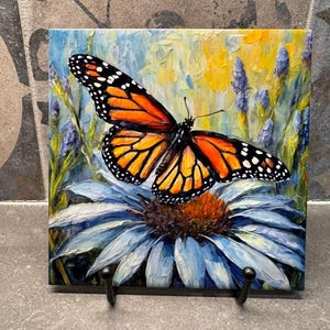 May include: Square ceramic tile featuring a vibrant painting of a monarch butterfly with orange and black wings, perched on a blue and orange flower. The background includes lavender and yellow brushstrokes, creating a colorful floral scene.