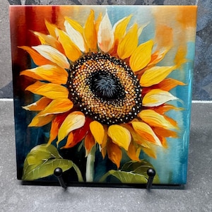 May include: A ceramic tile with a painted image of a sunflower with yellow petals and a dark brown center. The background is a blue and orange watercolor wash.