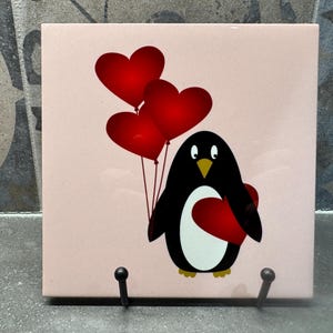 May include: A black and white penguin holding a red heart in its flippers. The penguin is standing on a pink background with three red heart-shaped balloons floating above it.