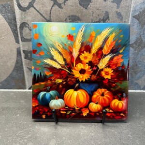 Puede incluir: Azulejo decorativo con una pintura de temática otoñal. La obra representa una naturaleza muerta con calabazas, girasoles y espigas de trigo en tonos naranja, amarillo y azul. El azulejo tiene dos ganchos negros.