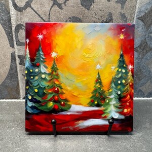 May include: A painting of a winter scene with five Christmas trees in a snowy forest. The trees are decorated with colorful ornaments and lights. The sky is a bright yellow and orange, with a few clouds. The painting is on a square tile with a black stand.