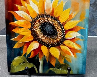 Pode incluir: Um azulejo de cerâmica com uma imagem pintada de um girassol com pétalas amarelas e um centro castanho escuro. O fundo é uma lavagem de aquarela azul e laranja.
