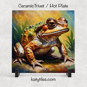 May include: Ceramic trivet featuring a detailed illustration of a frog with orange, green, and brown tones. The frog is set against a colorful, abstract background with green foliage. The text "Ceramic Trivet / Hot Plate" and "katytiles.com" are visible.