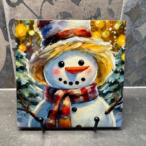 Puede incluir: Pintura cuadrada de un muñeco de nieve con nariz de zanahoria, ojos de botón negros y una bufanda a rayas rojas y blancas. El muñeco de nieve lleva un sombrero de paja y está sobre un fondo nevado con árboles y luces amarillas.