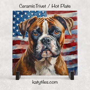May include: Square ceramic hot plate featuring a Boxer dog against a backdrop of the American flag. The dog has brown and white fur and a serious expression. The trivet rests on a black stand. Text at the top reads "Ceramic Trivet / Hot Plate".