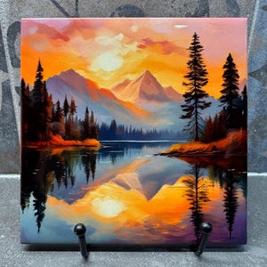 May include: Square wall art depicting a landscape painting of a mountain lake at sunset. The artwork shows a colourful sky with orange and yellow hues, reflected in the water. Silhouetted trees frame the scene. Includes two black hooks.