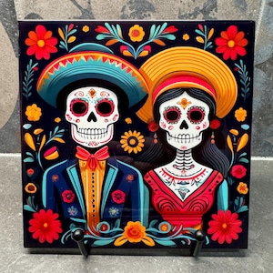 May include: A colorful ceramic tile with a Day of the Dead theme. The tile features two skeletons dressed in traditional Mexican clothing, with floral patterns and bright colors.