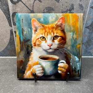 May include: A ceramic tile with a painting of an orange and white cat holding a blue and white coffee mug. The cat is looking directly at the viewer with a serious expression.