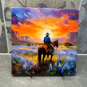May include: A square decorative tile featuring a cowboy on horseback silhouetted against a vibrant sunset. The artwork depicts a landscape with bluebonnets, mountains, and a lone tree. The tile has two black hooks attached to the bottom.