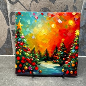 May include: A painting of a winter scene with three Christmas trees decorated with colorful ornaments. The trees are silhouetted against a bright orange, yellow, and blue sky. The painting is on a small canvas and is mounted on a black easel.