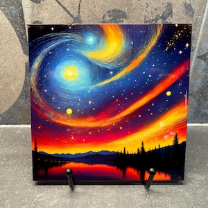 May include: Square artwork showing a colourful night sky with swirling galaxies in blue, yellow, and orange. The image includes a reflective lake and silhouetted trees, with a starry sky. The piece is displayed on a black metal stand.