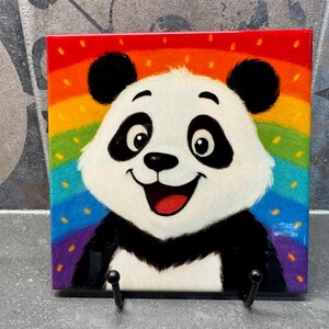 May include: A square decorative tile depicting a smiling panda bear set against a rainbow backdrop. The panda has black ears, eyes, and markings, with a white face and a red open mouth. The tile is mounted on a black metal stand.
