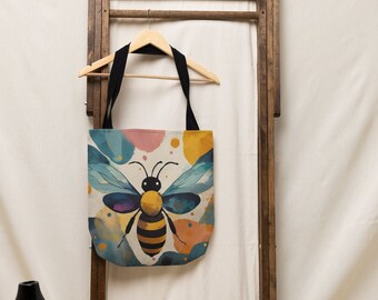 May include: A beige tote bag with black straps features a watercolor illustration of a bee with yellow and black stripes on its body. The bee is surrounded by colorful abstract shapes.