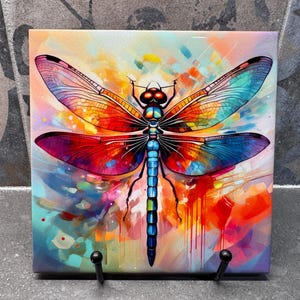 May include: A colorful square art print featuring a vibrant dragonfly illustration. The dragonfly has detailed wings in shades of orange, red, blue, and purple. The background is a blend of abstract colors, creating a striking visual.