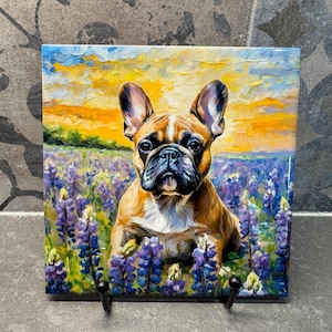 May include: Square ceramic tile featuring a French Bulldog in a field of purple flowers, with a sunset background. The dog has brown and white fur and is the focal point of the artwork. The tile is displayed on a black metal stand.