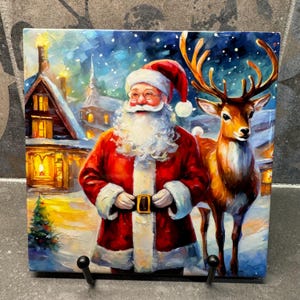 Puede incluir: Un azulejo de cerámica con temática navideña que presenta una pintura de Papá Noel con un traje rojo, barba blanca y un reno. El fondo es una escena invernal nevada con una casa y un árbol de Navidad.