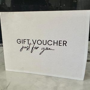 May include: A white gift voucher with the words "GIFT VOUCHER" in a bold, sans-serif font and "just for you..." in a cursive script. The voucher is rectangular and stands upright on a light-colored surface.