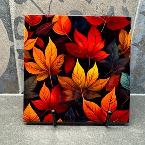 Puede incluir: Azulejo decorativo cuadrado con un vibrante diseño de hojas de otoño. Las hojas están representadas en tonos de rojo, naranja y amarillo sobre un fondo oscuro. El azulejo se exhibe sobre un soporte de metal negro.