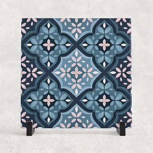 May include: A square decorative tile featuring a repeating floral pattern in shades of blue, navy, and pink. The tile is displayed on a black stand against a neutral background. The design includes stylized flowers and geometric shapes.