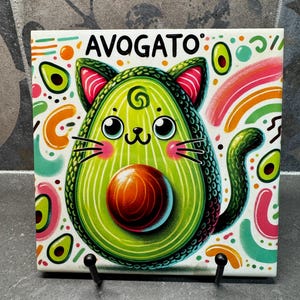 Può includere: Una piastrella di ceramica bianca con un'illustrazione colorata di un avocado a forma di gatto. L'avocado ha un guscio verde, un nocciolo marrone e un viso di gatto rosa e bianco. La piastrella ha la parola "AVOGATO" scritta in nero in alto.