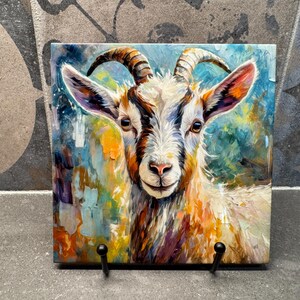 May include: A square ceramic tile featuring a colorful, impressionistic painting of a goat's face. The goat has white fur, brown horns, and is set against a backdrop of blues, greens, and oranges. The tile is displayed on a black metal stand.