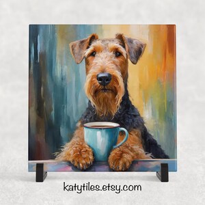 May include: A ceramic tile featuring a painted Airedale Terrier with a brown and black coat, holding a blue mug with a dark liquid. The background has a colorful, abstract design. The tile is displayed on a black stand. The Etsy shop URL is visible.