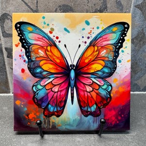 Pode incluir: Azulejo quadrado com uma ilustração vibrante de borboleta. As asas da borboleta exibem um espectro de cores, incluindo laranja, vermelho, azul e roxo, sobre um fundo abstrato e colorido. O azulejo é suportado por um suporte de metal preto.