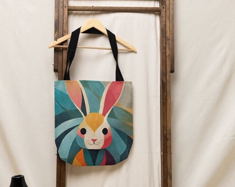 May include: A colorful tote bag with a geometric design of a rabbit's face. The rabbit has pink ears, a yellow face, and blue and green fur.