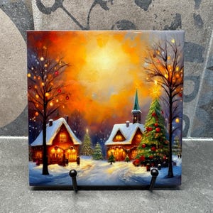 May include: A winter village scene with two snow-covered houses, a Christmas tree, and a bright orange sunset. The houses have glowing windows and the tree is decorated with red and gold ornaments.