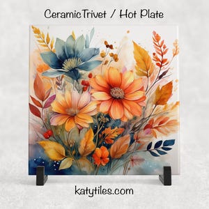 Puede incluir: Un salvamanteles o placa caliente de cerámica cuadrada con un diseño floral de acuarela. La obra de arte presenta flores naranjas y amarillas, flores azul-verdes y hojas de colores otoñales. El texto "Ceramic Trivet / Hot Plate" está en la parte superior.