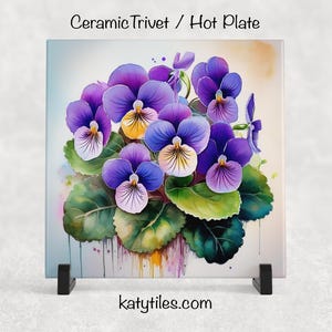 Puede incluir: Salvamanteles cuadrado de cerámica con una pintura de acuarela de pensamientos morados con centros amarillos y hojas verdes. El salvamanteles tiene el texto "Ceramic Trivet / Hot Plate" y "katytiles.com".