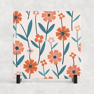 May include: A square ceramic coaster with a floral pattern. The coaster features a repeating pattern of orange and pink flowers with teal green stems and leaves on a light pink background.