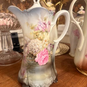 May include: A porcelain coffee pot featuring a scalloped rim, a curved handle, and a lid with a finial. The body is decorated with a floral arrangement of pink roses, white hydrangeas, and small yellow flowers against a cream and grey base.
