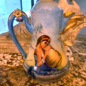 Op de afbeelding: Een porseleinen koffiepot met een lichtblauwe achtergrond en gouden accenten op het handvat en de deksel. De body toont een handgeschilderde scène van een persoon met een rode hoofdtooi en blauw kledingstuk, die een grote beige kruik vasthoudt. Signatuur Cairo.