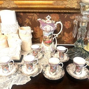 Puede incluir: Un juego de té de porcelana que incluye una jarra alta con tapa y seis tazas a juego con platillos. Cada pieza muestra una colorida escena pastoral con figuras, acentuada por adornos morados y dorados sobre una base blanca con bordes.
