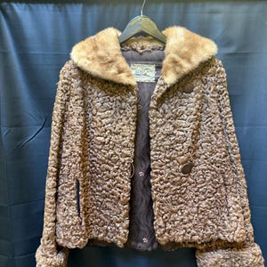 May include: A brown, textured jacket with a fur collar and cuffs. The jacket has a button closure and a dark lining. The label reads "46335". The jacket is hanging on a wooden hanger against a dark blue backdrop.