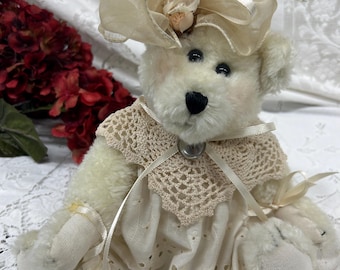 May include: A vintage-style teddy bear with a cream-colored plush body, wearing a cream-colored dress with eyelet details and a crocheted collar. The bear has a matching hat with a small rose and ribbon accents.