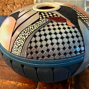 Puede incluir: Un jarrón decorativo de cerámica con un diseño geométrico único. El jarrón presenta una variedad de colores, incluyendo negro, gris, melocotón y blanco, con un patrón de tablero de ajedrez y texturas punteadas. La parte superior tiene una abertura circular.