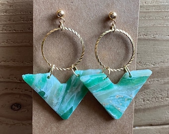 May include: A pair of green and gold earrings with a geometric chevron shape. The earrings are made of resin and have a marbled effect. The gold hoops are attached to the top of the chevron shape.